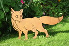 A metal garden stake shaped like a fox, made from rusted orange Corten steel, standing on a green lawn in front of leafy plants.