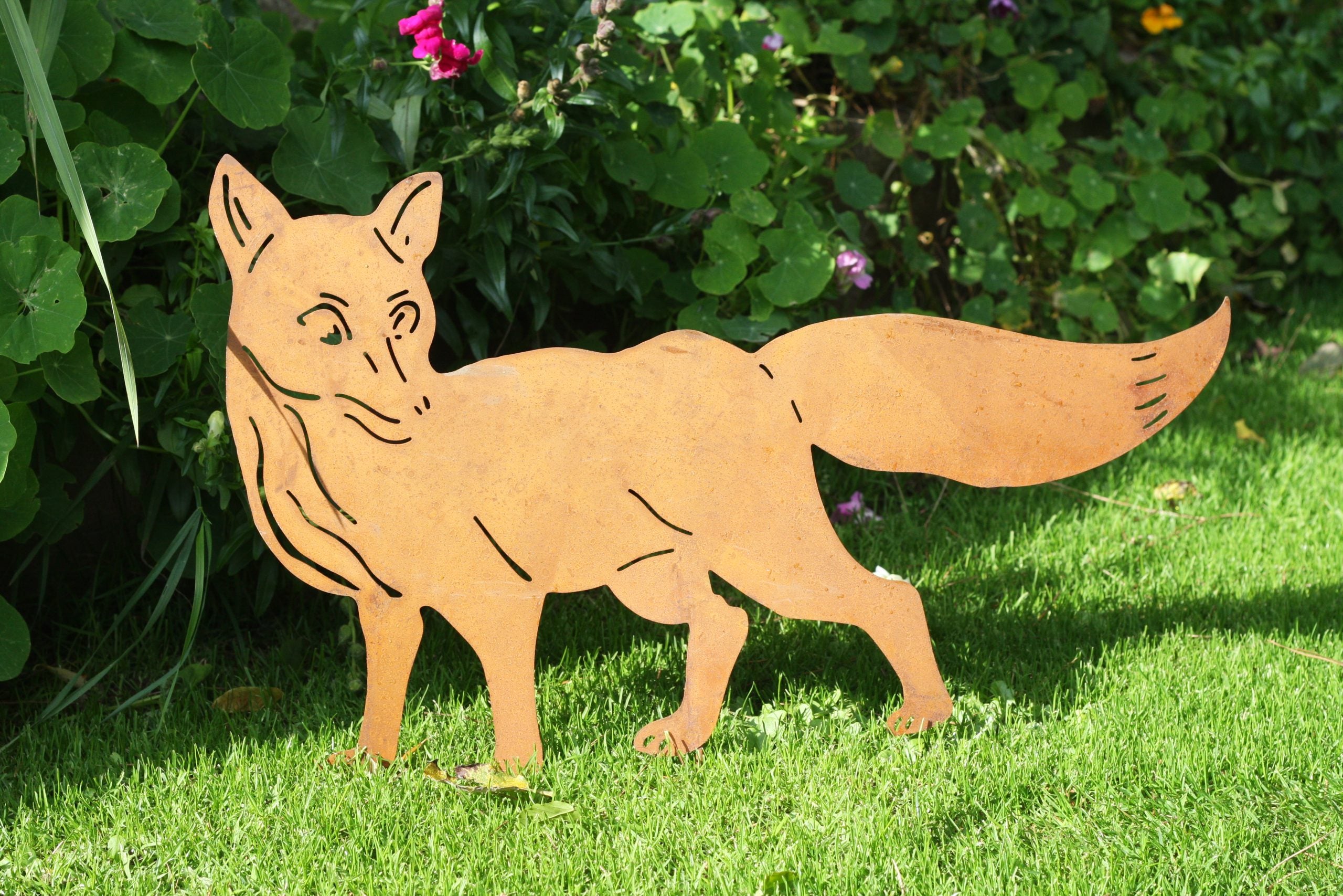 A metal garden stake shaped like a fox, made from rusted orange Corten steel, standing on a green lawn in front of leafy plants.