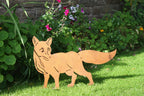 A metal garden cutout of a fox standing on green grass in front of a stone wall and leafy plants with pink and purple flowers.