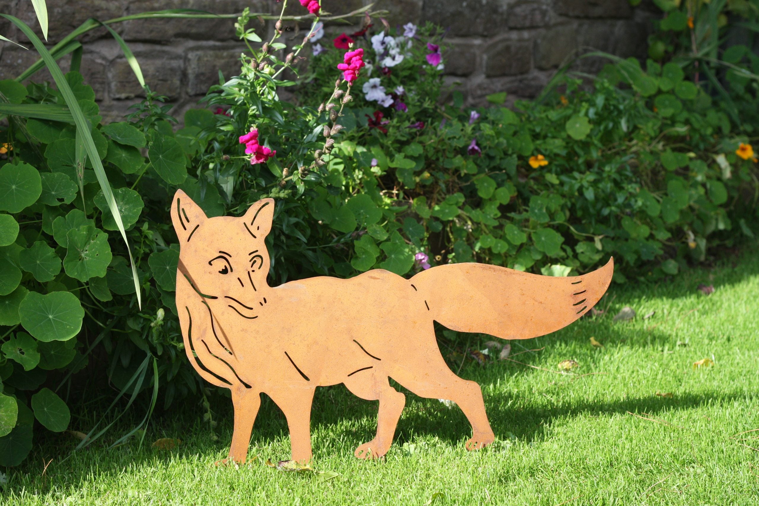 A metal garden cutout of a fox standing on green grass in front of a stone wall and leafy plants with pink and purple flowers.