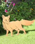 A light brown wooden fox silhouette yard sign standing on a green lawn in front of a stone wall and garden plants.