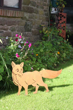 A light brown wooden fox silhouette yard sign standing on a green lawn in front of a stone wall and garden plants.