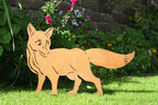 A wooden fox yard stake shaped like a walking fox, standing on a green lawn in front of a garden wall with plants and flowers.