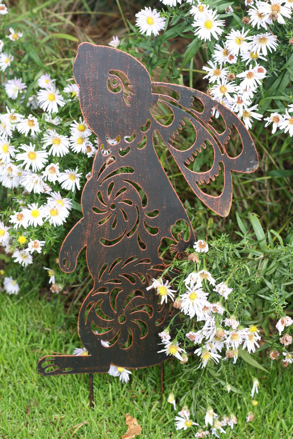A dark brown metal garden stake shaped like a fairy with intricate cutout patterns, displayed in a garden setting among green grass and white daisies.