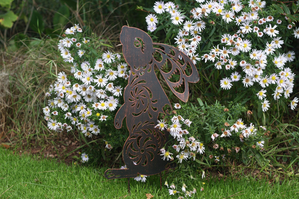 A dark brown metal garden stake shaped like a rabbit with decorative cutout details, placed in a grassy garden bed surrounded by white daisies.