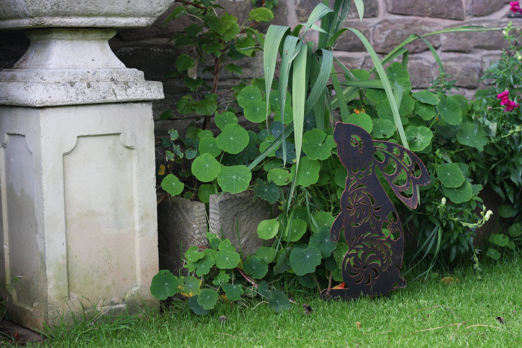 A dark metal garden angel cutout with intricate scrollwork, displayed outdoors on a green lawn next to a stone wall and potted plants.