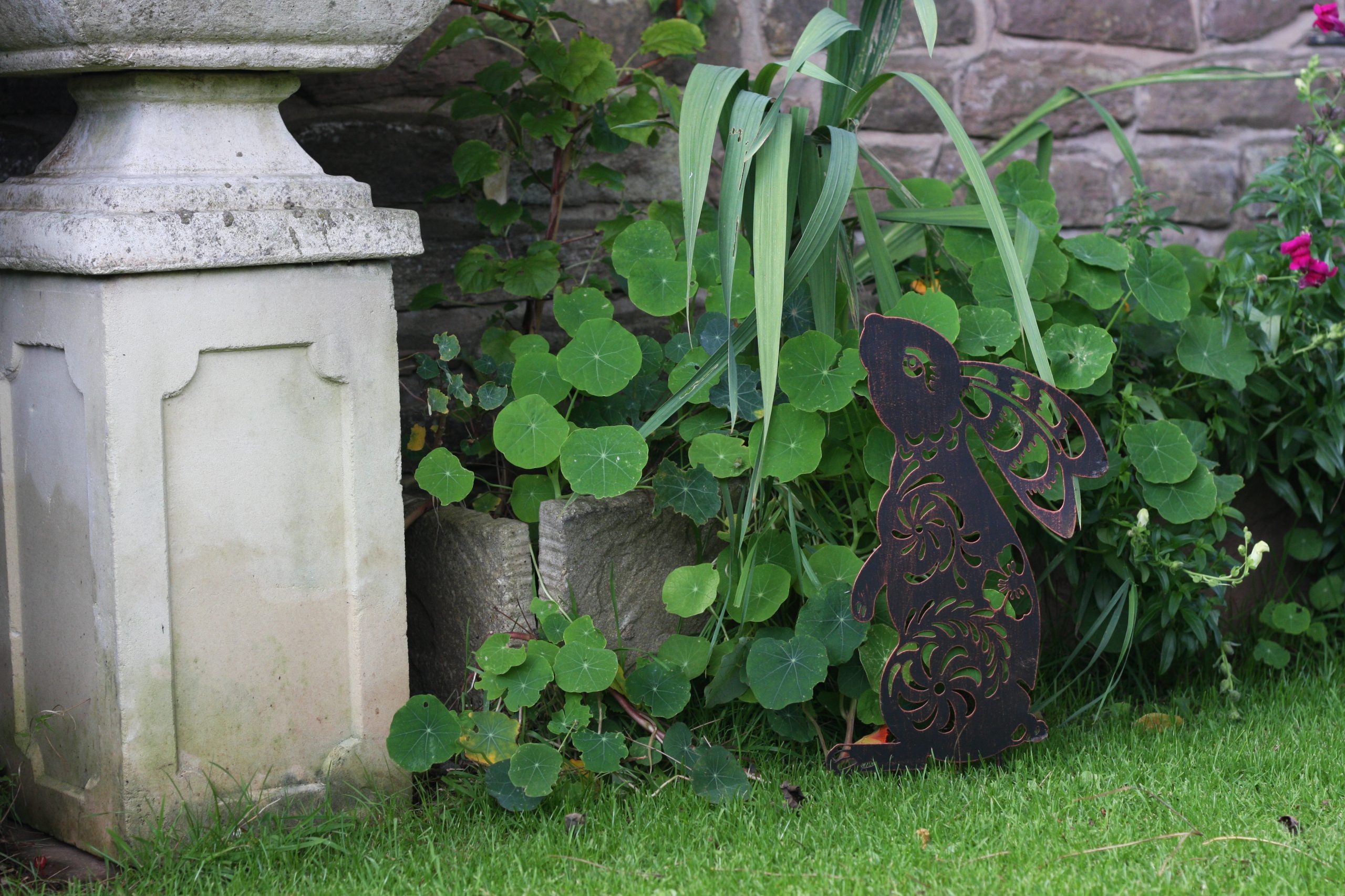 A dark metal garden angel cutout with intricate scrollwork, displayed outdoors on a green lawn next to a stone wall and potted plants.