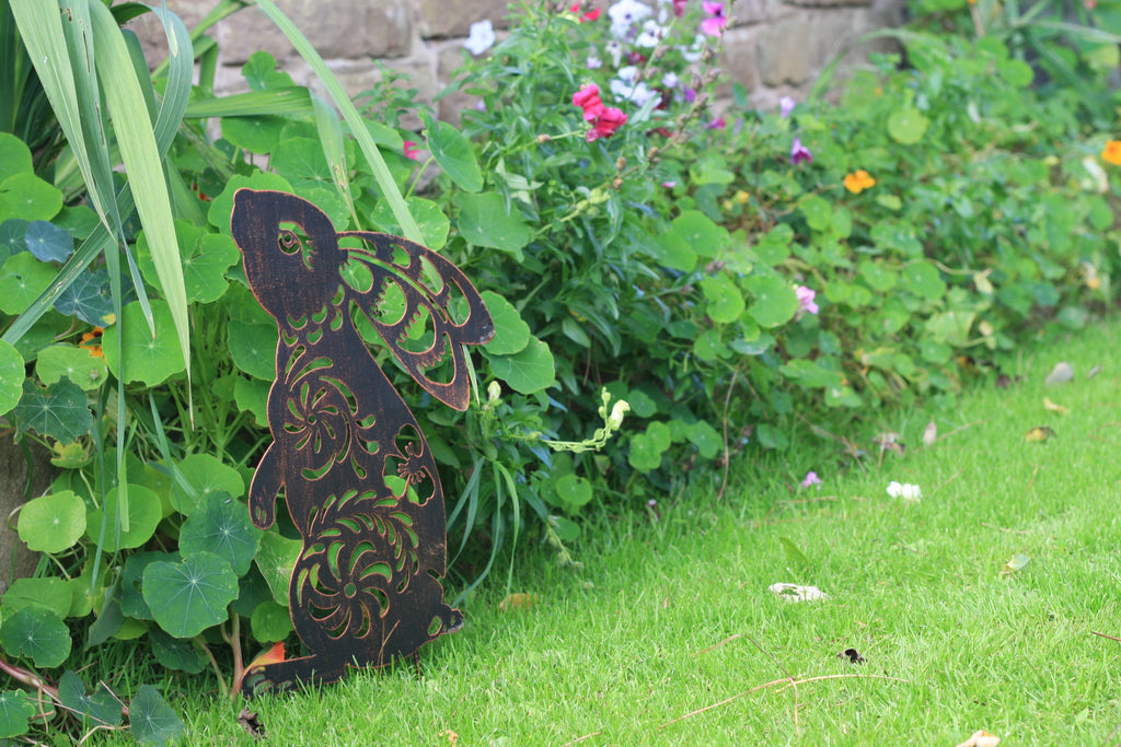 A dark brown wooden angel garden stake with intricate cutout patterns, standing on a green lawn next to lush garden plants and flowers.