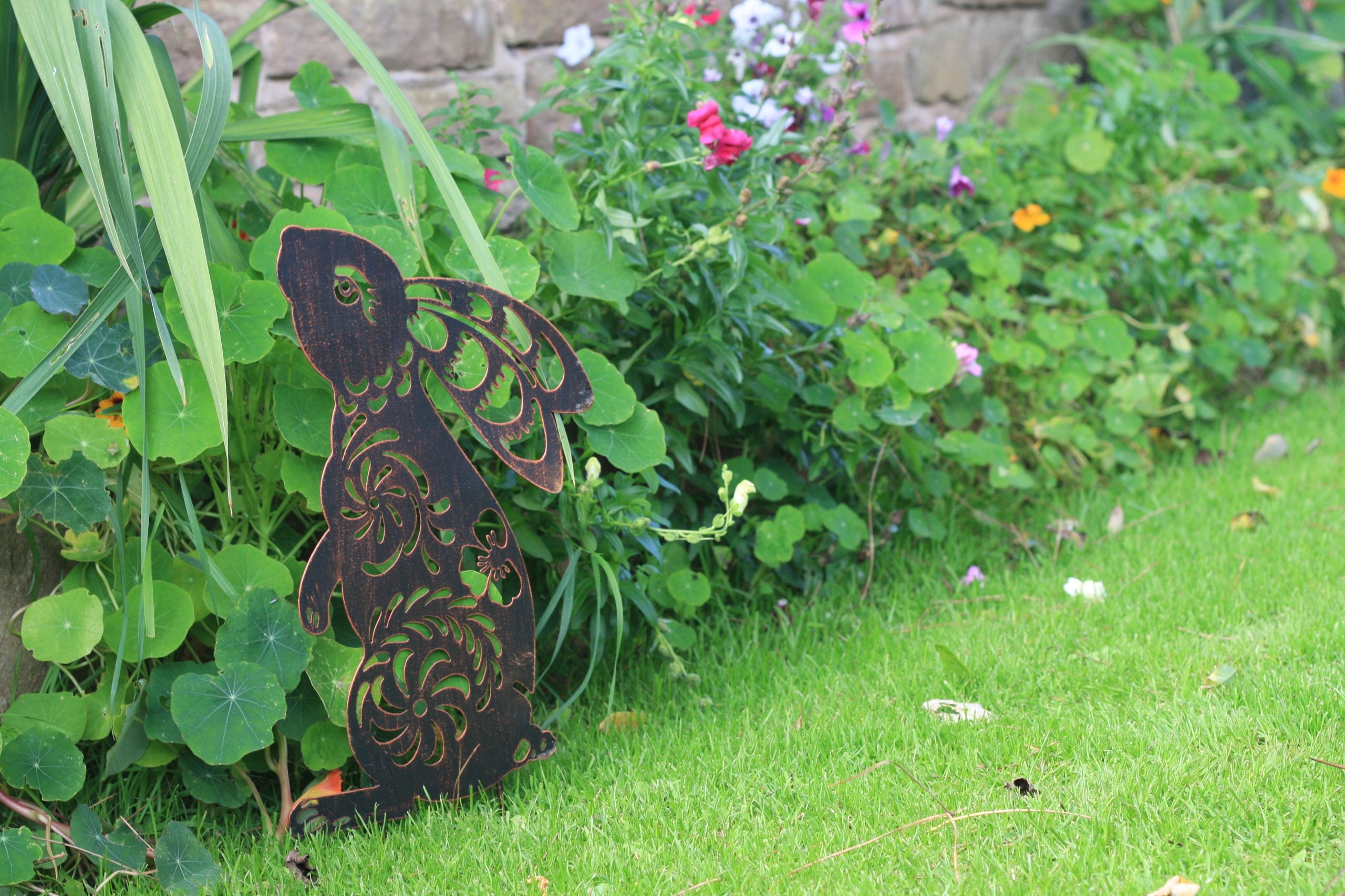 A dark brown wooden angel garden stake with intricate cutout patterns, standing on a green lawn next to lush garden plants and flowers.