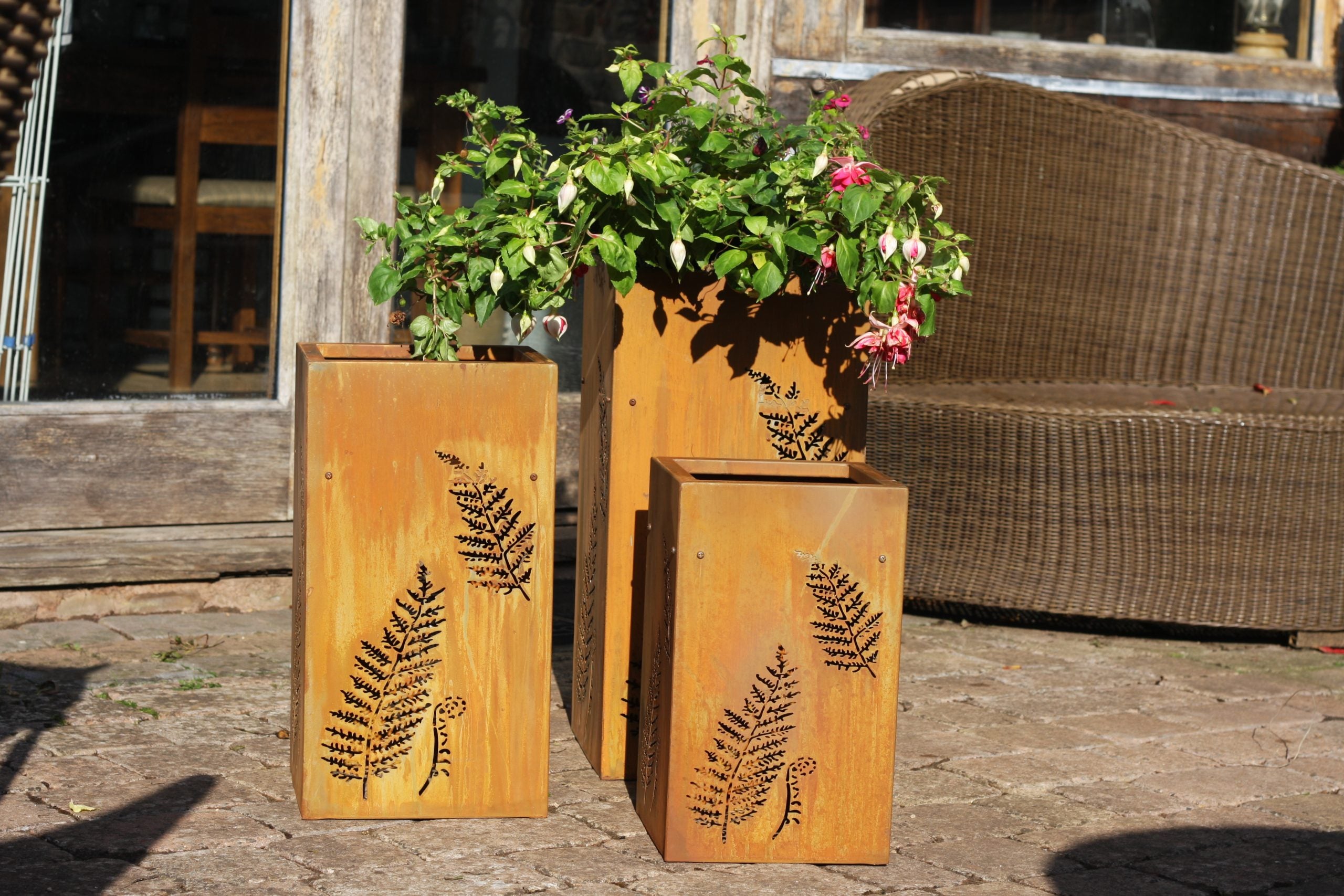 Three rectangular wooden planters with laser-cut fern patterns, arranged outdoors on a stone surface with a wicker chair and building in the background.