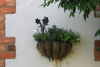 A rustic half-hoop coconut fiber hanging planter with a black metal flower accent, filled with green and purple flowers, mounted on a white stucco wall with red brick corners.