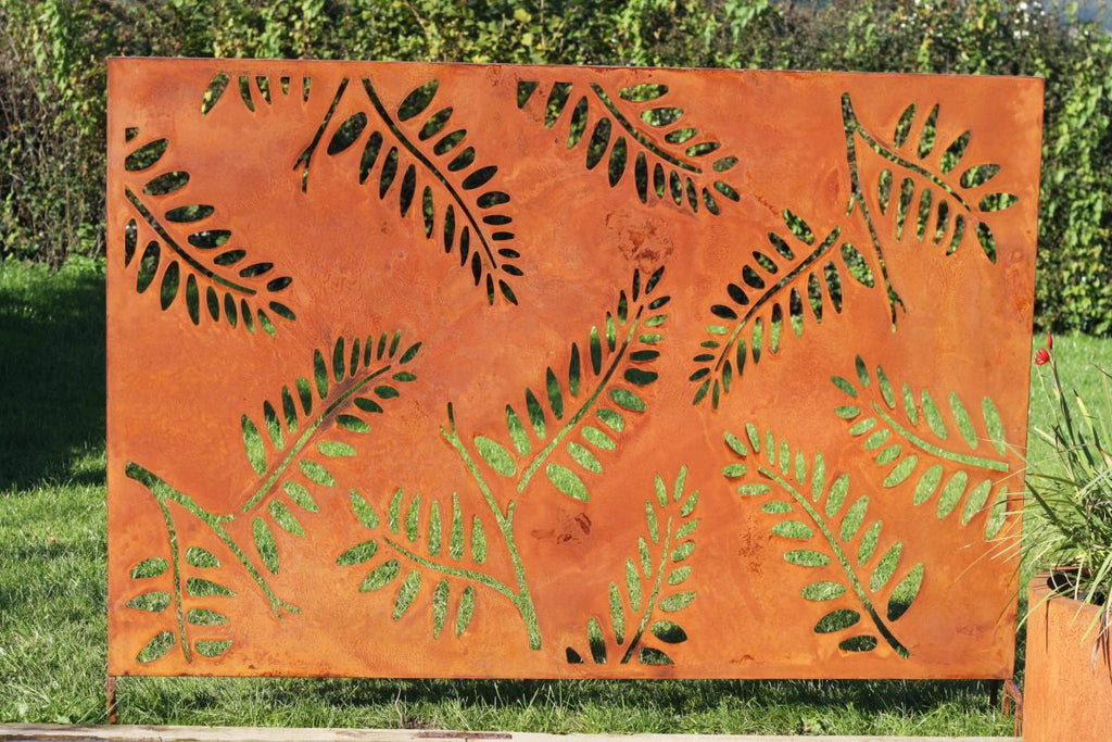 An orange rectangular metal garden sculpture panel with laser-cut green fern leaf patterns, displayed outdoors on a grassy lawn with green foliage in the background.