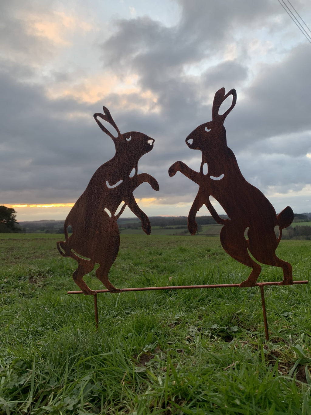 Two rusted metal hares facing each other on a wire stand in a grassy field under a cloudy sky.