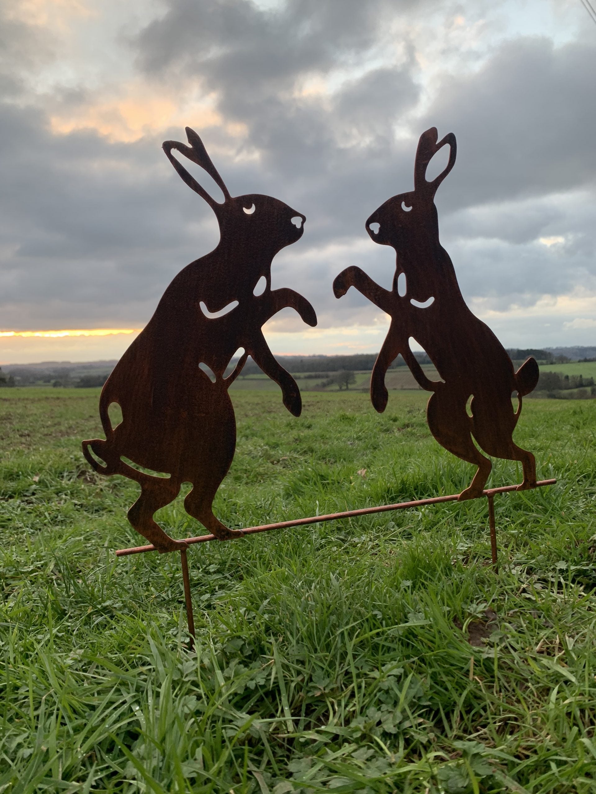 Two metal Easter bunnies standing on a garden stake in a grassy field under a cloudy sky.