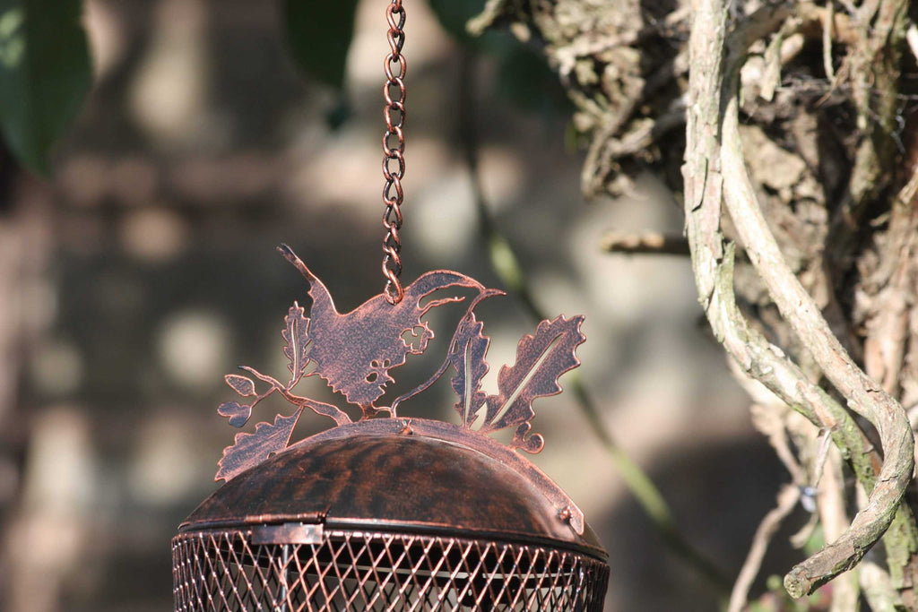 A rustic metal bird feeder hanging from a chain outdoors, featuring a wire mesh body and a decorative metal top shaped like leaves and a bird, shown against a blurred natural background.