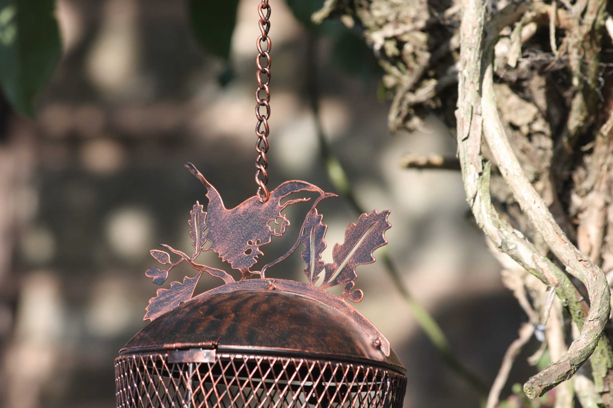 A rustic metal bird feeder hanging from a chain outdoors, featuring a wire mesh body and a decorative metal top shaped like leaves and a bird, shown against a blurred natural background.