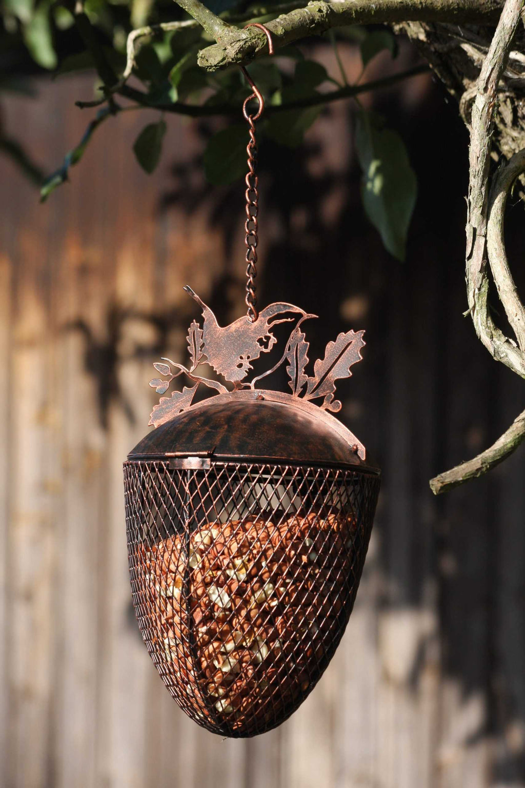 A copper acorn-shaped hanging bird feeder filled with seeds, suspended from a tree branch outdoors against a blurred wooden fence background.