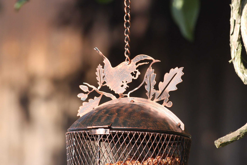 A close-up of a hanging metal bird feeder with a decorative bird silhouette on top, made from copper-toned metal with a dark patina, suspended by a chain outdoors.