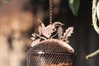 A close-up of a hanging metal bird feeder with a decorative bird silhouette on top, made from copper-toned metal with a dark patina, suspended by a chain outdoors.