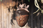 A metal bird feeder shaped like an acorn hangs from a chain outdoors. The feeder has a mesh body filled with seeds and a decorative metal top featuring a bird silhouette and leaf details, set against a blurred wooden fence background.