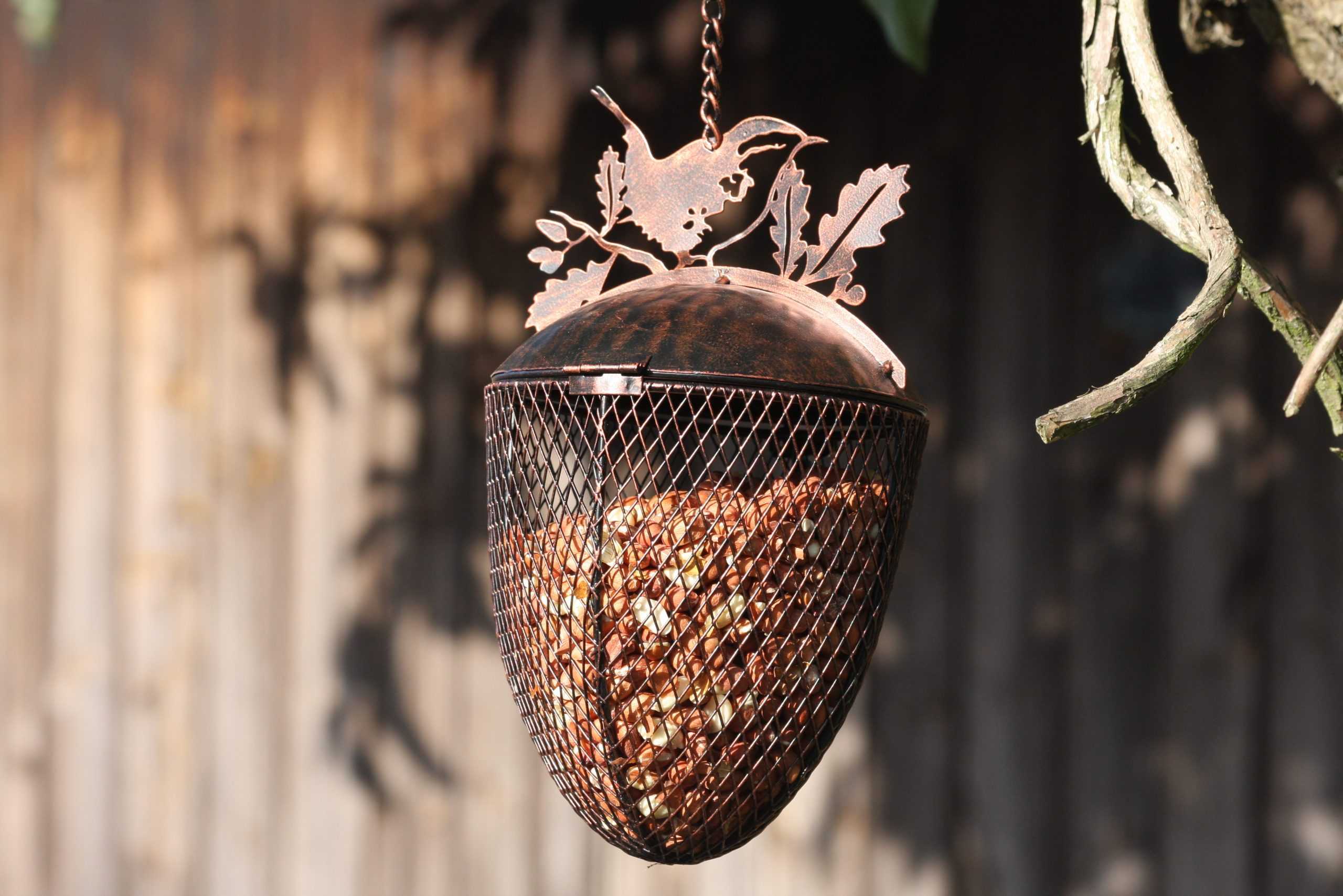 A metal bird feeder shaped like an acorn hangs from a chain outdoors. The feeder has a mesh body filled with seeds and a decorative metal top featuring a bird silhouette and leaf details, set against a blurred wooden fence background.