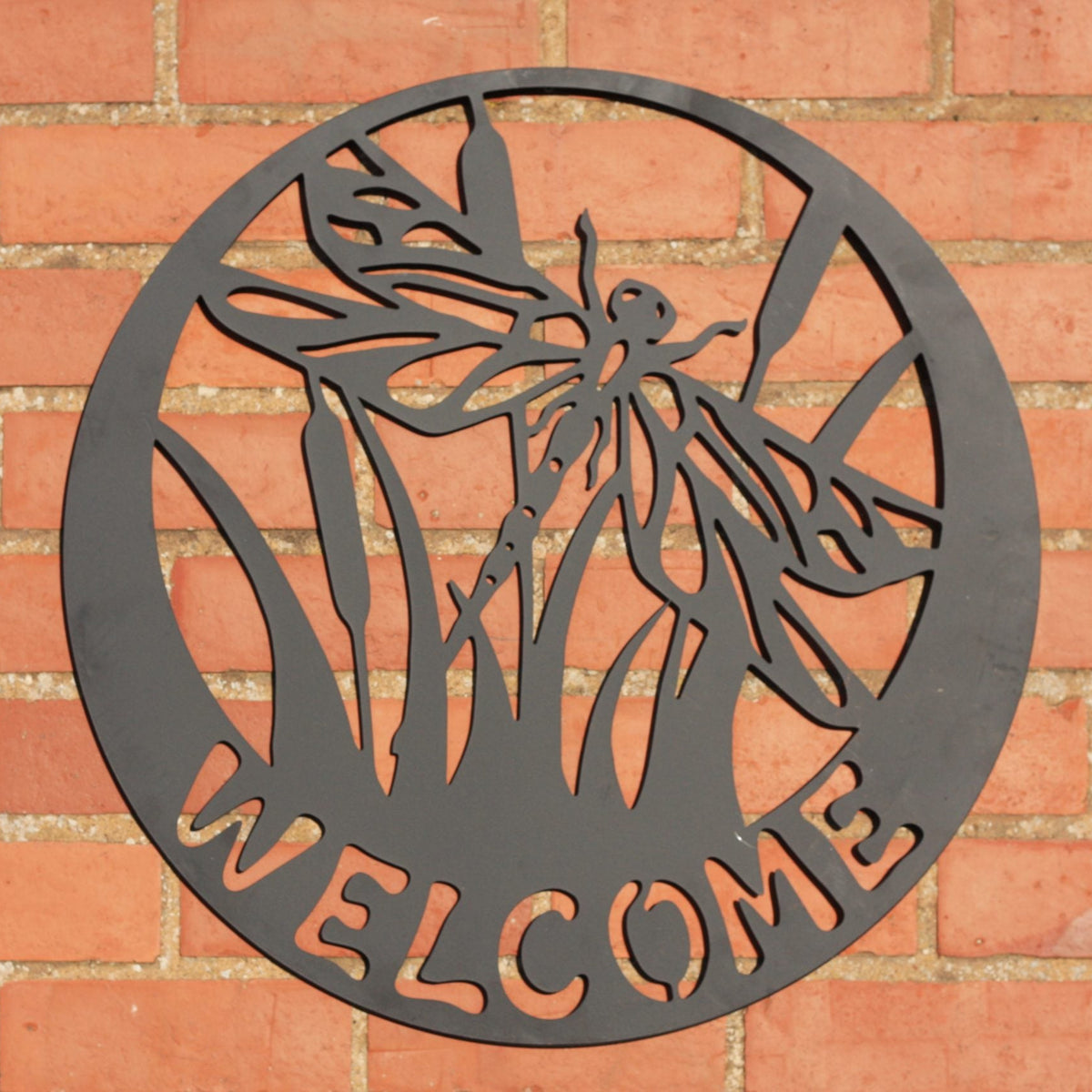 A circular black metal wall sign featuring a dragonfly silhouette and the word 'WELCOME' in cutout letters, mounted on a red brick wall.