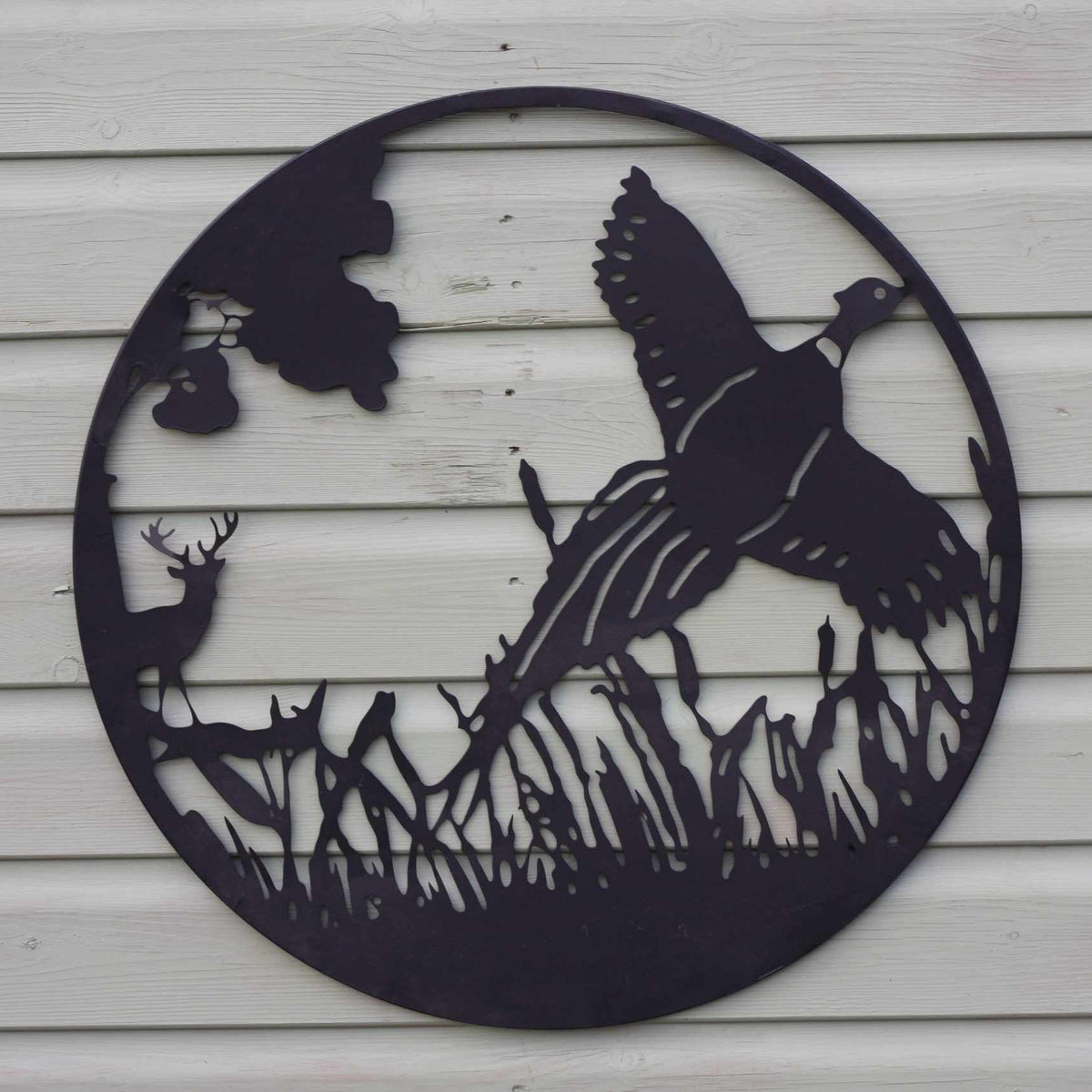 A circular black metal wall art piece featuring a pheasant, deer, duck, and reeds, mounted on a white horizontal siding wall.