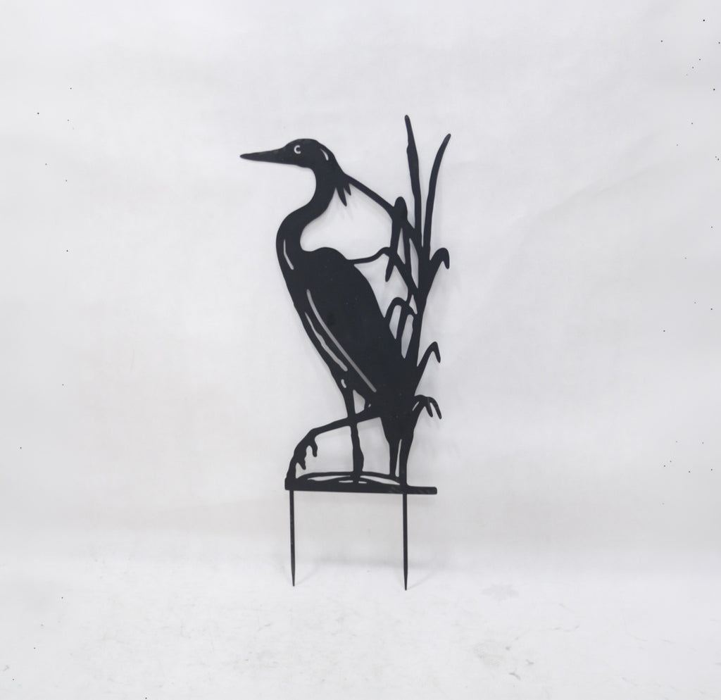 Black metal yard sign of a heron standing among reeds on a small stake, photographed against a plain light background.
