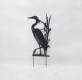 Black metal yard sign of a heron standing among reeds on a small stake, photographed against a plain light background.