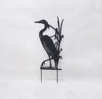 Black metal yard sign of a heron standing among reeds on a small stake, photographed against a plain light background.