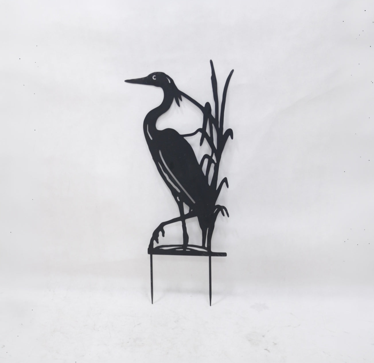 Black metal yard sign of a heron standing among reeds on a small stake, photographed against a plain light background.