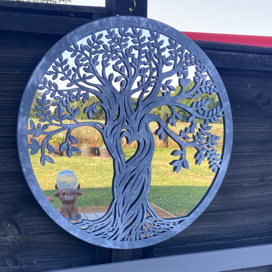 A circular metal garden wall art piece featuring a tree of life design, mounted on a dark wooden fence. The tree is cut out to show a view of a green lawn and a small dog statue inside, with a small bird figurine placed on the left side of the tree trunk.