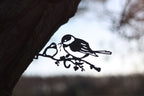 Black metal bird perched on a branch wall hanging, shown against a blurred outdoor background with tree bark and sky.