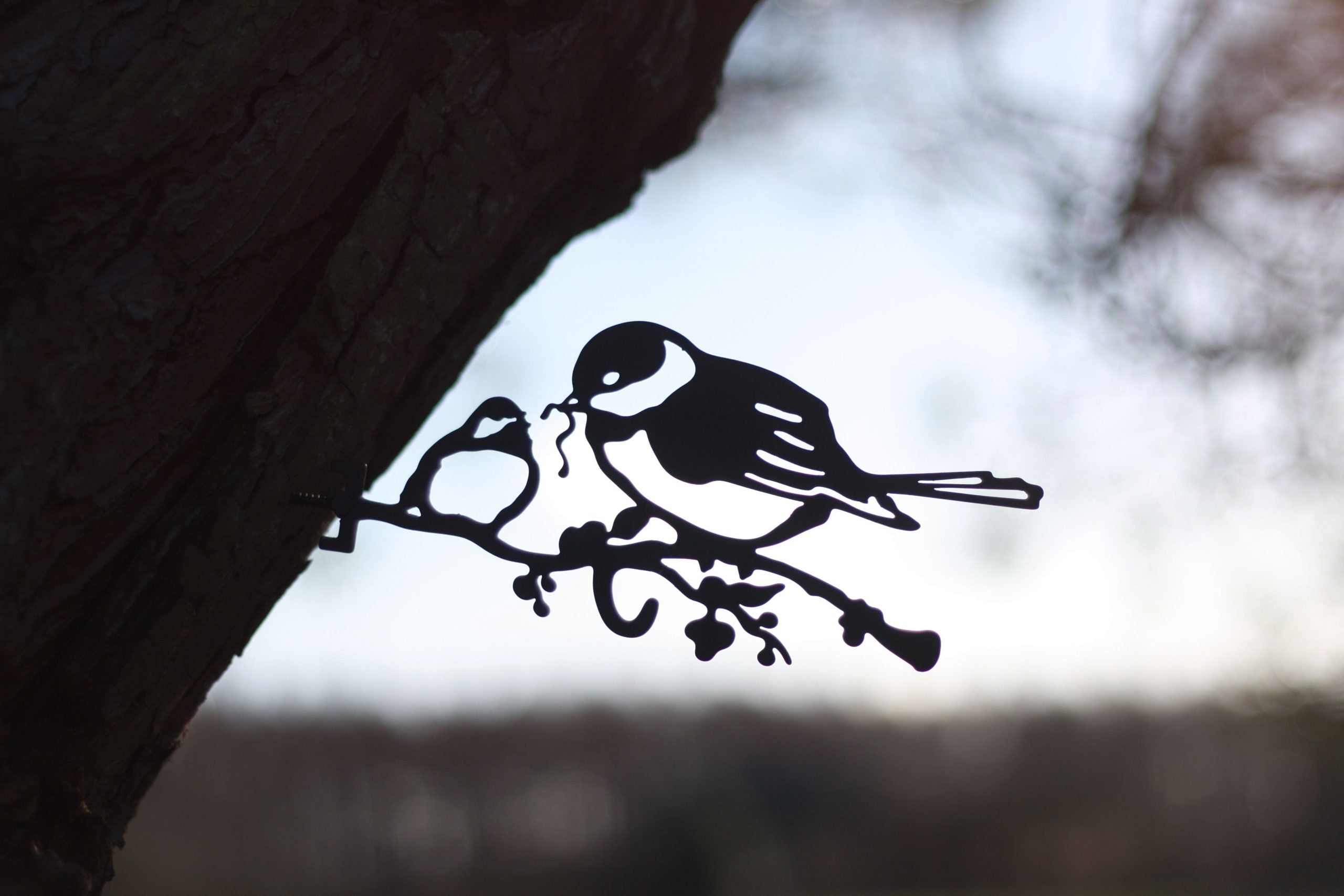 Black metal bird perched on a branch wall hanging, shown against a blurred outdoor background with tree bark and sky.