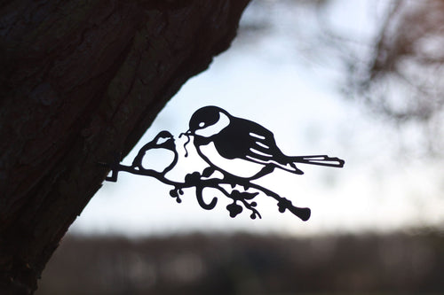 Black metal bird perched on a branch wall hanging, shown against a blurred outdoor background with tree bark and sky.