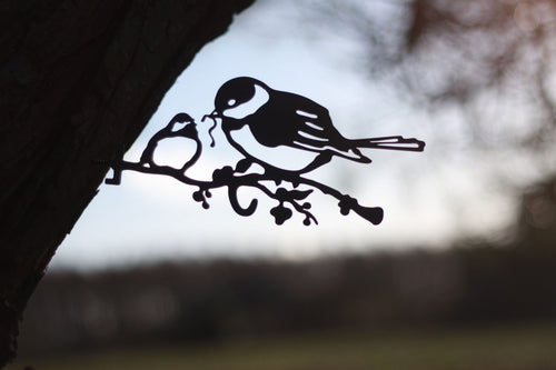 Black metal bird perched on a branch garden sign, shown in silhouette against a blurred outdoor background.