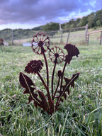 Rustic metal garden sculpture of dandelions and leaves in a dark reddish-brown finish, displayed in a green grassy outdoor garden setting under a cloudy sky.