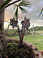 Two dark metal garden stake flower sculptures shaped like ornamental daisies, planted in a rustic stone planter outdoors with a blurred grassy yard and cloudy sky in the background.