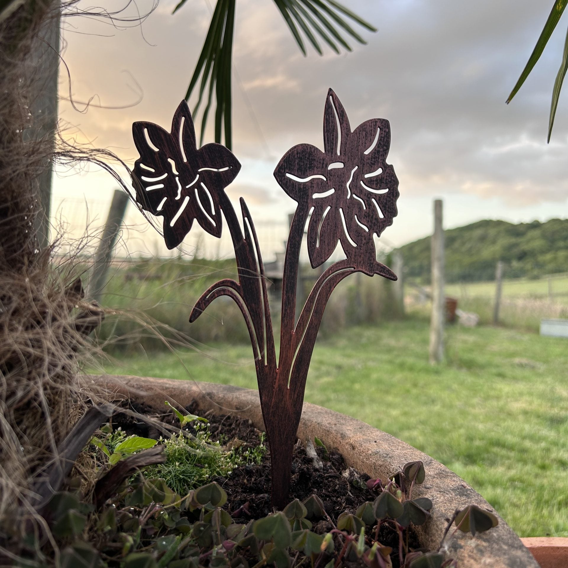 Two dark metal daffodil garden stakes planted in a rustic stone planter outdoors, with a blurred grassy field and trees in the background.