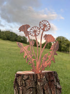 A copper-colored metal dandelion garden sculpture displayed on a rustic tree stump in an outdoor grassy field under a cloudy sky.