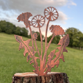 Copper metal dandelion garden stake mounted on a rustic tree stump, photographed outdoors against a blurred green lawn and blue sky.
