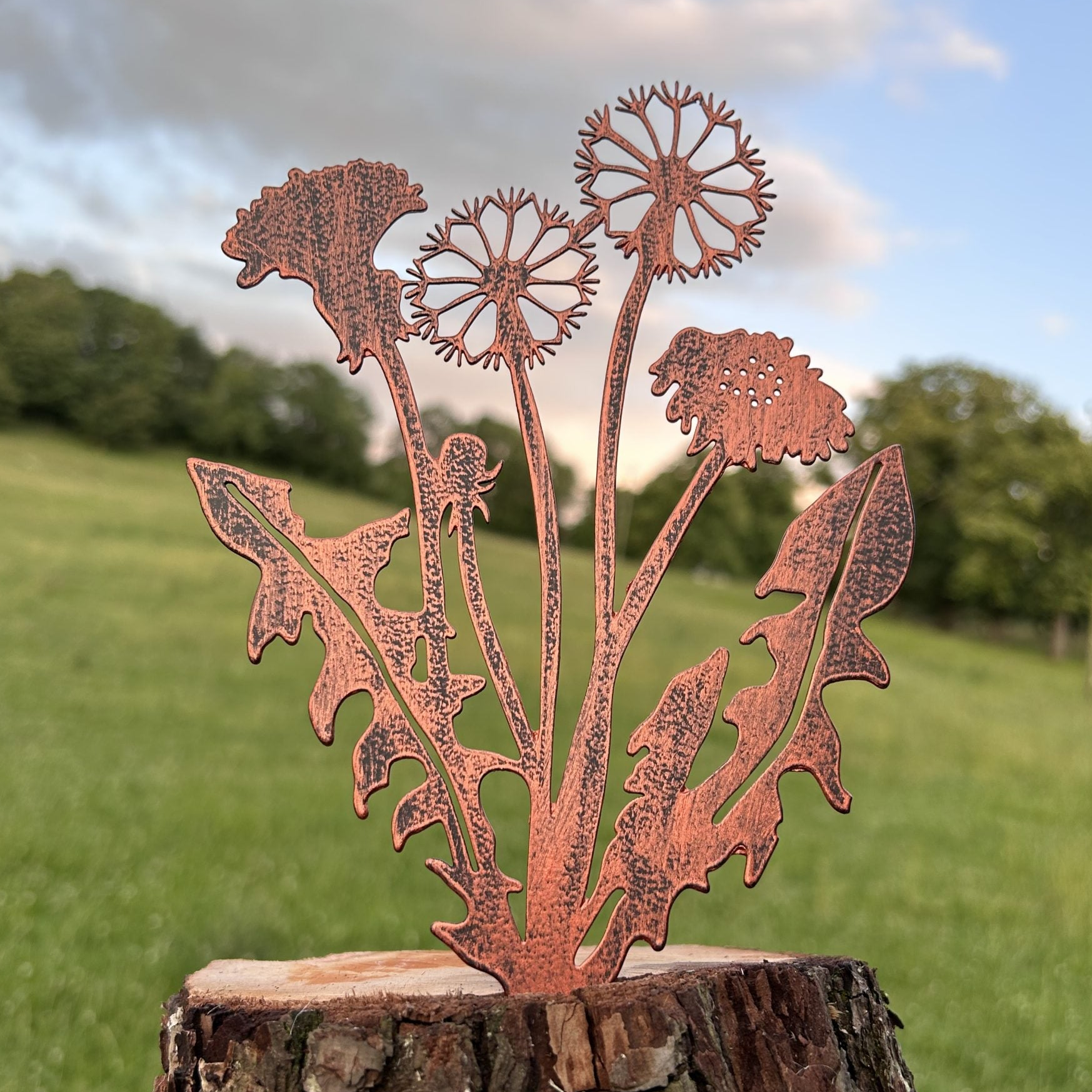 Copper metal dandelion garden stake mounted on a rustic tree stump, photographed outdoors against a blurred green lawn and blue sky.