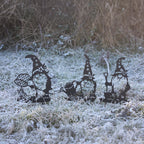 Three black metal garden gnome cutouts with winter-themed accessories, displayed on frosty grass outdoors.