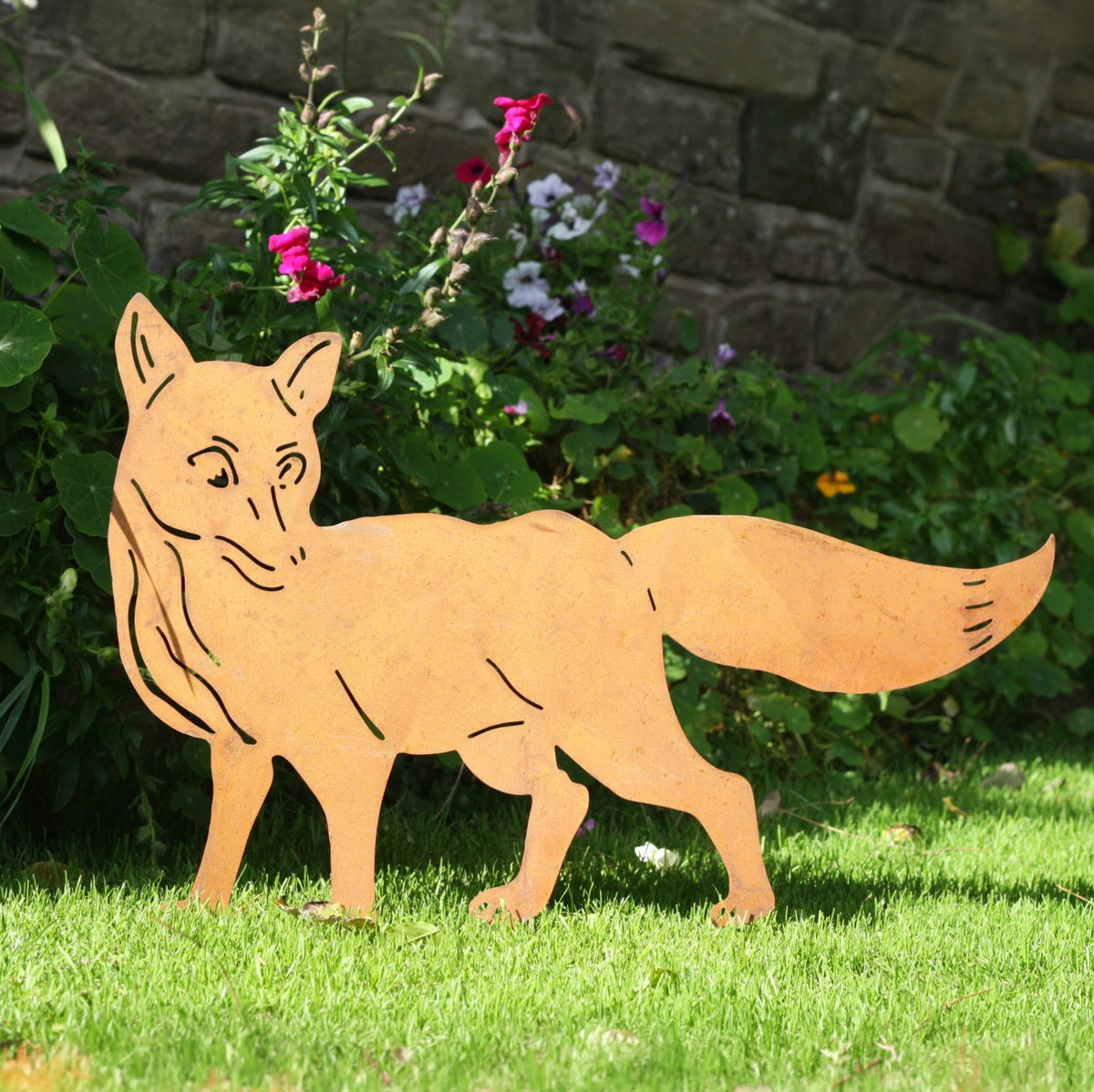 A laser-cut metal garden stake shaped like a fox standing on a green lawn in front of a stone wall and garden plants.