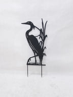 Black metal garden stake featuring a heron standing among reeds, mounted on two thin stakes against a plain white background.