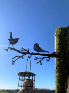 Black metal bird feeder and perch mounted on a moss-covered wooden post outdoors under a clear blue sky, with two birds silhouetted on the branch and a hanging bird feeder below.