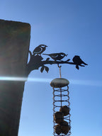A black metal bird feeder mounted on a tree branch, featuring silhouettes of three birds perched on the branch above a hanging seed basket, photographed against a clear blue sky.