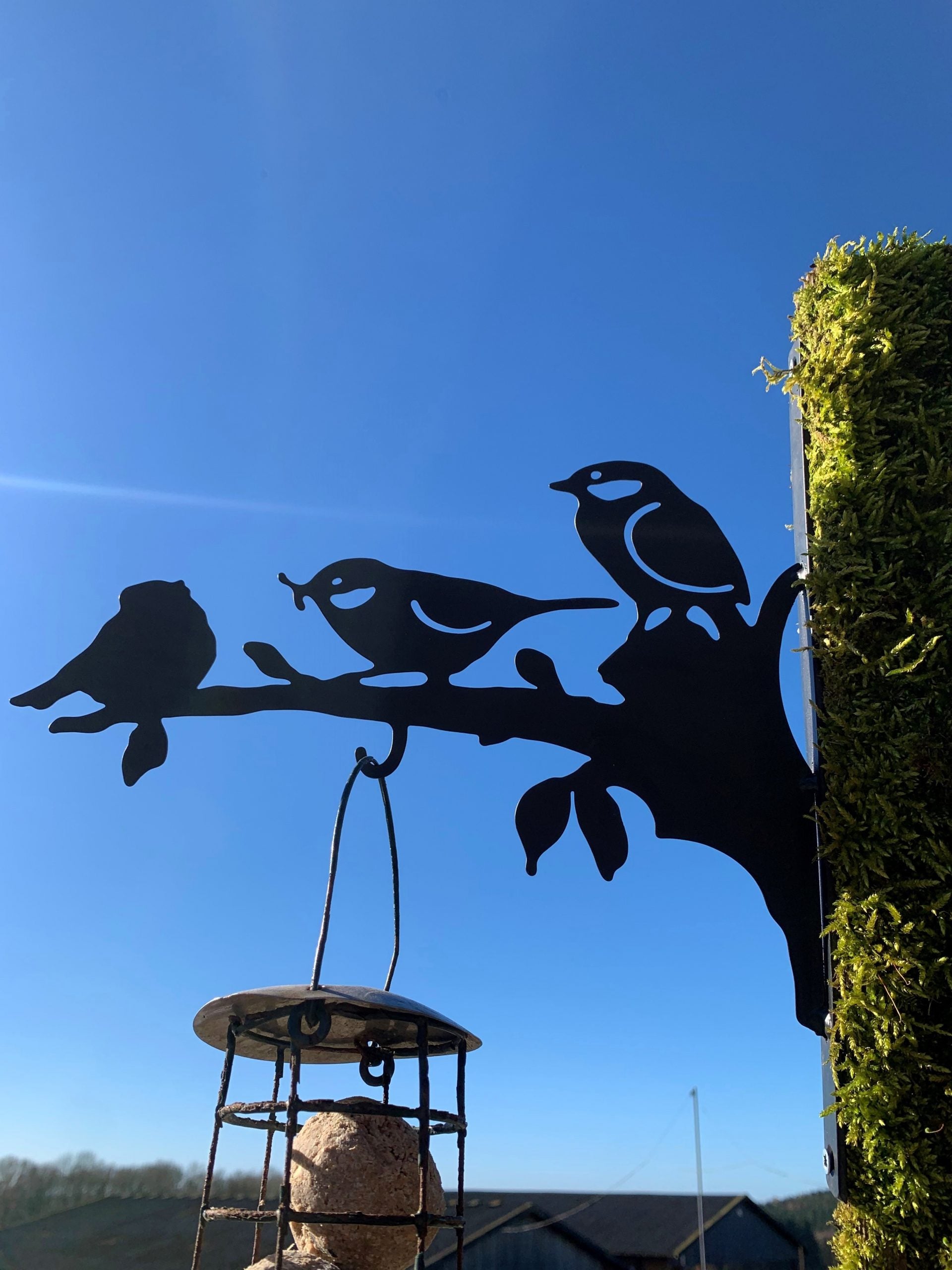 Black metal bird feeder shaped like a branch with three birds perched on it, hanging against a clear blue sky with a moss-covered post beside it.